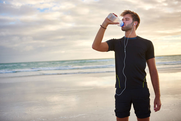 Νεαρός άνδρας φαίνεται σε φόρμα και σπορ πόσιμο από το μπουκάλι του νερού, ενώ απολαμβάνοντας ένα πρωινό τρέξιμο στην παραλία - Φωτογραφία, εικόνα
