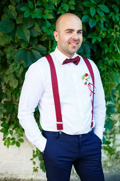 Portret van een man bruidegom met een rode vlinderdas en bretels. Hij is zelfverzekerd met een glimlach op zijn gezicht. Stijlvolle modieuze man met baard in een wit shirt en broek. - Foto, afbeelding