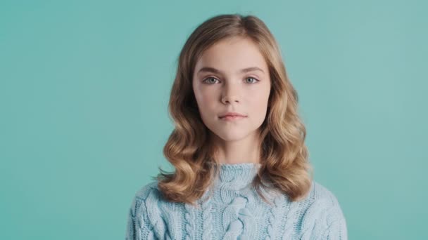 Menina adolescente muito loira vestida com suéter aconchegante olhando atentamente na câmera sobre fundo azul. Expressão facial - Filmagem, Vídeo