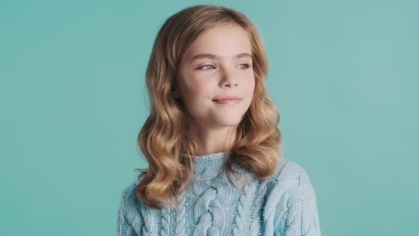 Beautiful blond teenage girl dressed in cozy sweater posing on camera isolated on blue background. Young model in studio - Footage, Video