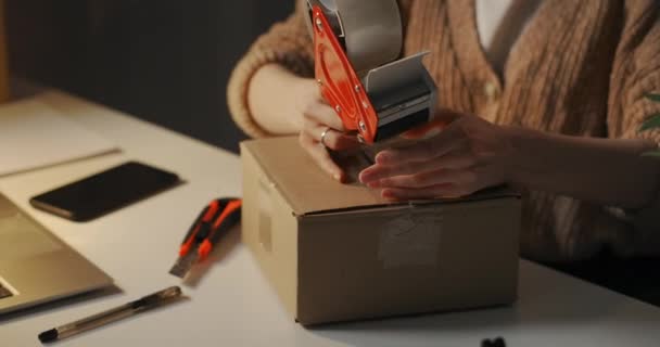 γυναίκα σφραγίζει κουτί από χαρτόνι με roll-on συσκευή διανομής ταινία στο τραπέζι εργασίας της το βράδυ, υπηρεσία παράδοσης - Πλάνα, βίντεο