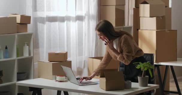 busy woman is talking with clients by mobile phone and checking information in laptop, working in home warehouse - Footage, Video