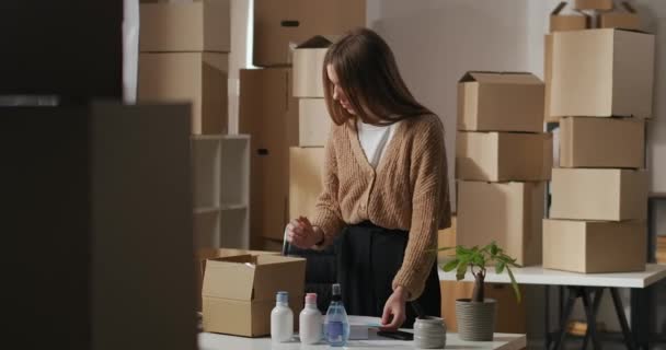 ενήλικη γυναίκα αποσυσκευασία κουτί με καλλυντικά σε μικρή αποθήκη στο διαμέρισμα, τον έλεγχο της παραγγελίας στο έγγραφο, τις μικρές επιχειρήσεις και την επιχειρηματικότητα - Πλάνα, βίντεο