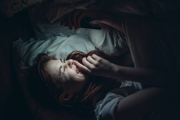 Ellerinde telefon olan duygusal bir kadın gece bağımlılığında yatakta yatıyor. - Fotoğraf, Görsel