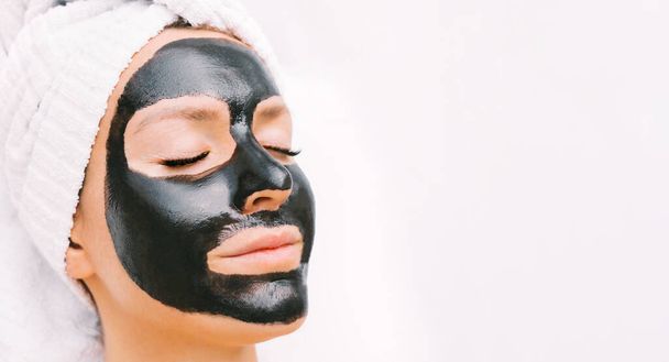 Siyah kömür temizleyici yüz maskeli güzel bir genç kadın. Kozmetik prosedürler, kaplıca salonunda cilt bakımı maskesi. Boşluğu kopyala - Fotoğraf, Görsel
