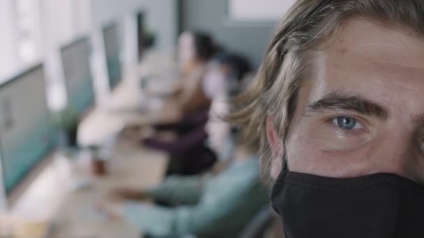 Extreme close-up met PAN van de man in het gezicht masker te kijken naar de camera in het kantoor, terwijl zijn onherkenbare collega 's werken aan computers op de achtergrond - Video