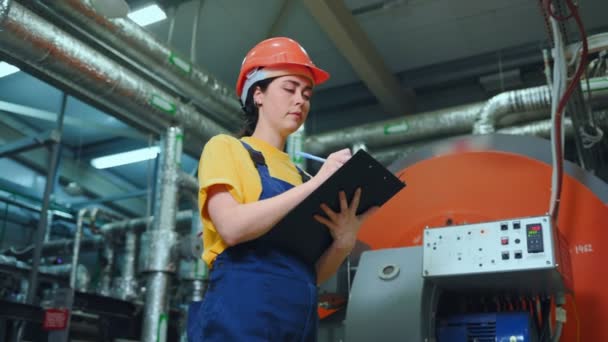 Nuori naisinsinööri seisoo tuotantohuoneessa ja kirjaa teknisten laitteiden lukemat. Alhaalta päin. Moderni teollisuus ja valmistus - Materiaali, video