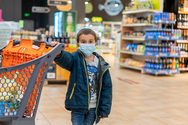 Il ragazzo indossa una maschera protetta in negozio. Tempo di shopping durante l'epidemia di coronavirus. Un ragazzo con una maschera medica. Shopping con i bambini durante l'epidemia di virus. Ragazzo in maschera in supermercato - Foto, immagini