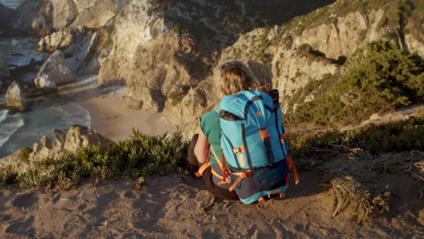 Vista trasera de la mujer con mochila sentada en el acantilado - Imágenes, Vídeo