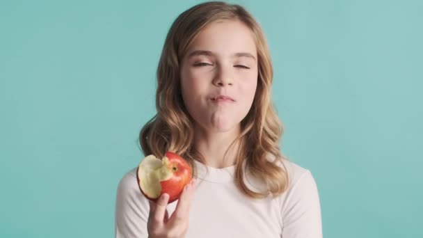 Vonzó szőke tinédzser lány eszik finom alma tartja hüvelykujját a kamera elszigetelt kék háttérrel. Ideje harapni valamit. Tetszik a kifejezés. - Felvétel, videó