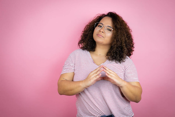 Mladá Afroameričanka v červených proužcích trička přes růžové pozadí s rukama u sebe a prsty zkřížené úsměv uvolněně a vesele. Úspěch a optimismus - Fotografie, Obrázek
