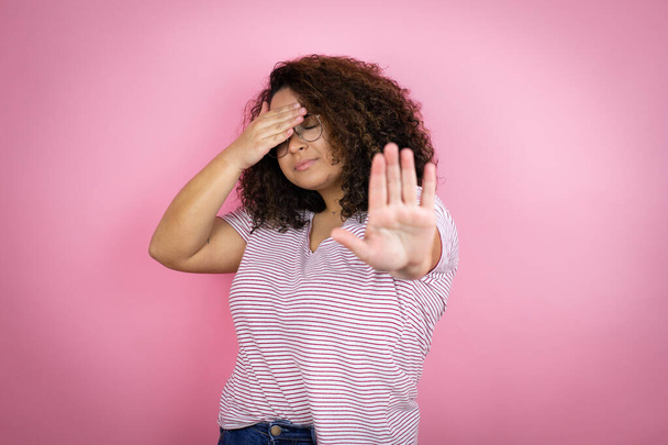 Молода афроамериканська жінка, одягнена в червону футболку над рожевим фоном, закриває очі руками і робить стоп жест з сумним і страшним виразом. Засмучена і негативна концепція. - Фото, зображення