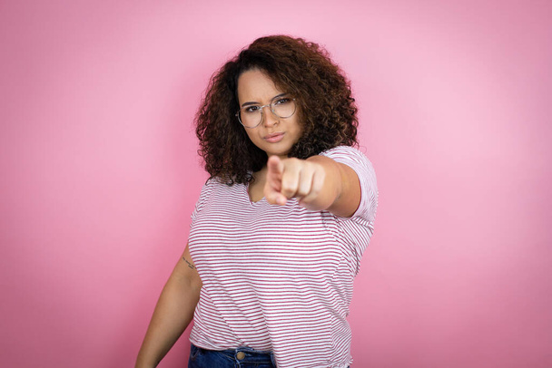 Mladá africká Američanka v červených pruhovaných tričkách na růžovém pozadí ukazující prstem na fotoaparát a na vás, sebevědomé gesto vypadá vážně - Fotografie, Obrázek