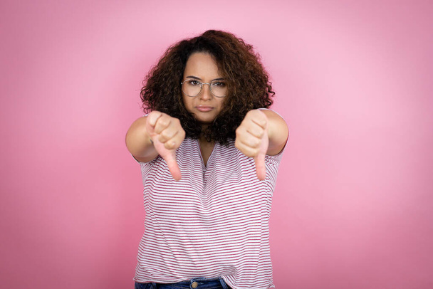 Young african american woman wearing red stripes t-shirt over pink background with angry face, negative sign showing dislike with thumbs down - Photo, Image