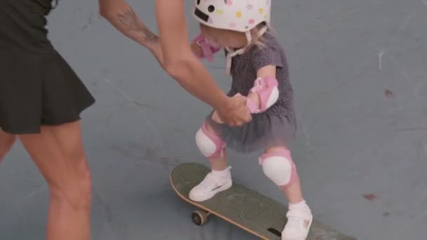 Vista posteriore della giovane donna caucasica che si tiene per mano la sua piccola figlia indossando casco di sicurezza colorato e pastiglie e aiutando il suo skateboard - Filmati, video