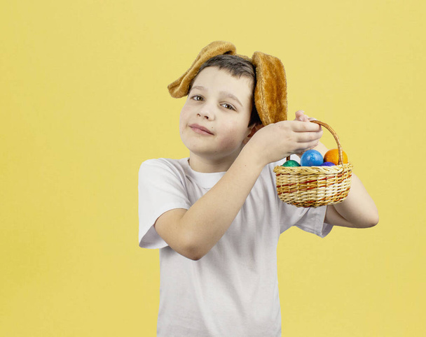 Αγόρι με αυγά. Κουνέλι στο κεφάλι του αγοριού. Πασχαλινό λαγουδάκι και καλάθι με αυγά σε κίτρινο φόντο. Πασχαλινά αυγά. Χαρούμενο Πάσχα. Πολύχρωμα πασχαλινά αυγά. Τα παιδιά γιορτάζουν το Πάσχα. Αντιγραφικός χώρος - Φωτογραφία, εικόνα