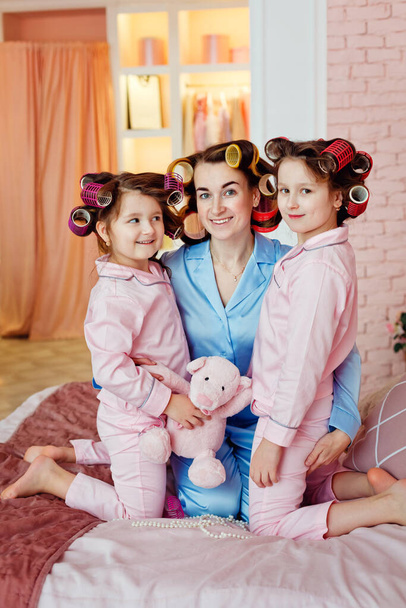 Mama i dziewczynki siedzą na łóżku w różowych lokach i niebieskiej piżamie. Piżama party. - Zdjęcie, obraz