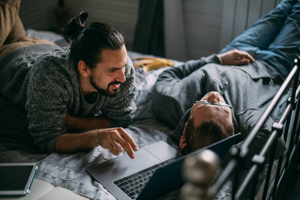 Młodzi mężczyźni leżą w łóżku z laptopem w domu. Przystojni, zrelaksowani mężczyźni surfujący po sieci i rozmawiający w sypialni na wygodnym łóżku w ciągu dnia - Zdjęcie, obraz