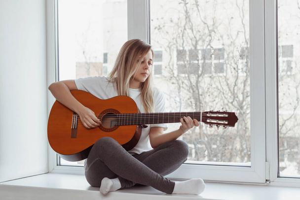 A young woman in the white t-shirt and grey leggings is sitting on the windowsill and playing acoustic guitar. Girl picks a chord clamping frets on the fretboard. Self-isolation. Lock down. Introvert - Foto, Bild