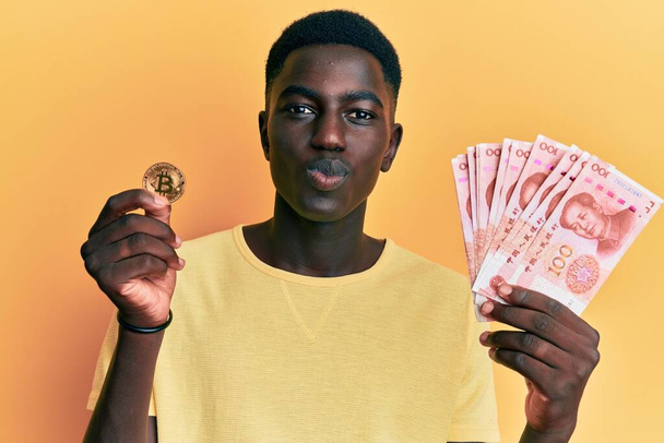 Young african american man holding yuan chinese banknotes and bitcoin looking at the camera blowing a kiss being lovely and sexy. love expression.  - Foto, Imagem