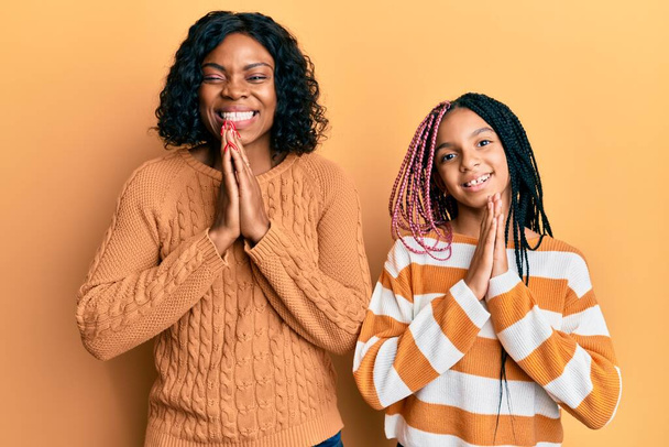 Schöne afrikanisch-amerikanische Mutter und Tochter tragen wollenen Winterpullover und beten mit den Händen zusammen um Vergebung und lächeln zuversichtlich.  - Foto, Bild