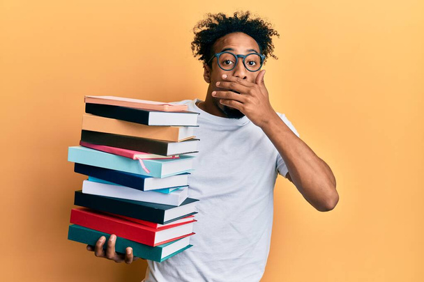 Young african american man with beard holding a pile of books covering mouth with hand, shocked and afraid for mistake. surprised expression  - Foto, Imagen