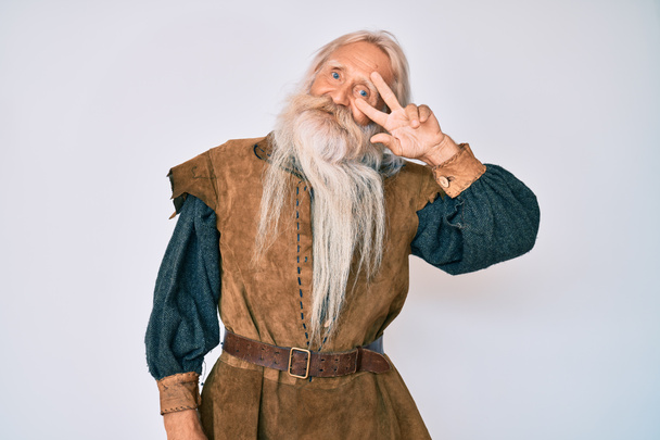 Vieil homme âgé aux cheveux gris et à la barbe longue portant un costume traditionnel viking faisant symbole de paix avec les doigts sur le visage, souriant et joyeux montrant la victoire  - Photo, image