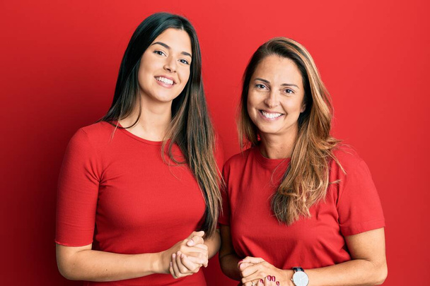 Spanyol család anya és lánya viselnek alkalmi ruhák felett piros háttér kezét össze, és keresztbe ujjak mosolyogva nyugodt és vidám. siker és optimizmus  - Fotó, kép