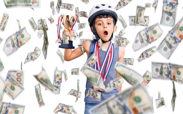 Pequeño niño lindo con casco de bicicleta y medallas ganadoras sosteniendo trofeo ganador asustado y sorprendido con la boca abierta para sorpresa, cara de incredulidad - Foto, imagen