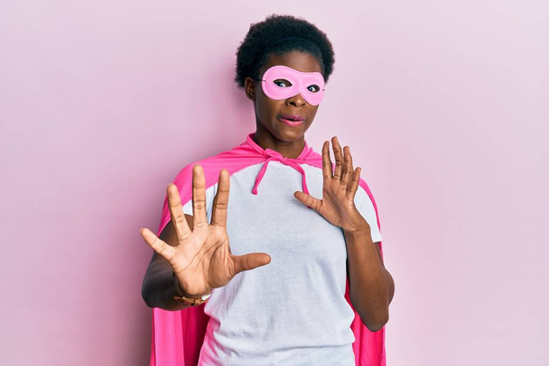 Young african american girl wearing superhero mask and cape costume afraid and terrified with fear expression stop gesture with hands, shouting in shock. panic concept.  - 写真・画像