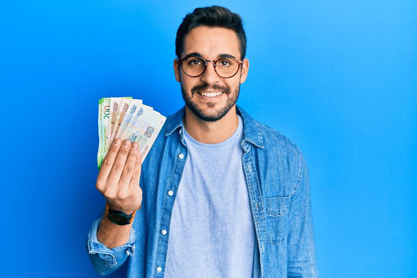 Joven hispano sosteniendo billetes de rublo rusos que lucen positivos y felices de pie y sonriendo con una sonrisa confiada mostrando los dientes  - Foto, imagen