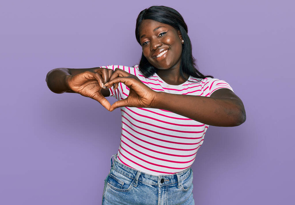 Африканская молодая женщина в повседневной полосатой футболке, улыбаясь в любви, делает символическую форму сердца руками. романтическая концепция.  - Фото, изображение