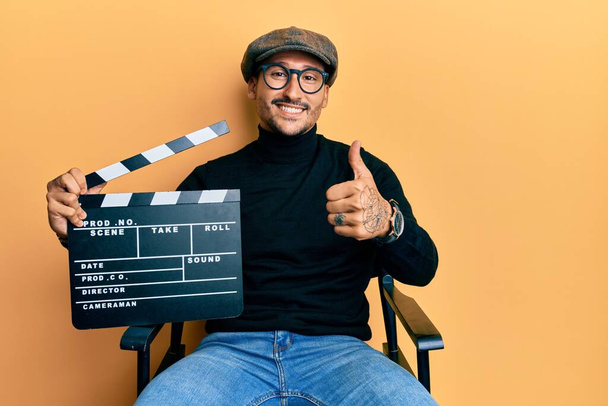 Handsome man with tattoos holding video film clapboard sitting on director chair smiling happy and positive, thumb up doing excellent and approval sign  - Photo, Image