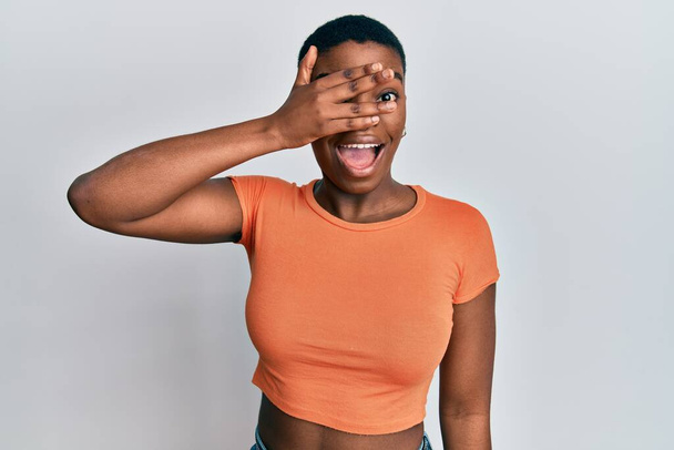 Fiatal afro-amerikai nő visel alkalmi narancssárga póló kukucskál sokk borító arc és a szemek kézzel, nézi az ujjakon keresztül zavarba ejtő kifejezés.  - Fotó, kép