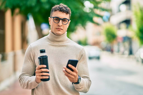 Young hispanic man wearing glasses using smartphone and holding bottle of water at the city. - Photo, Image