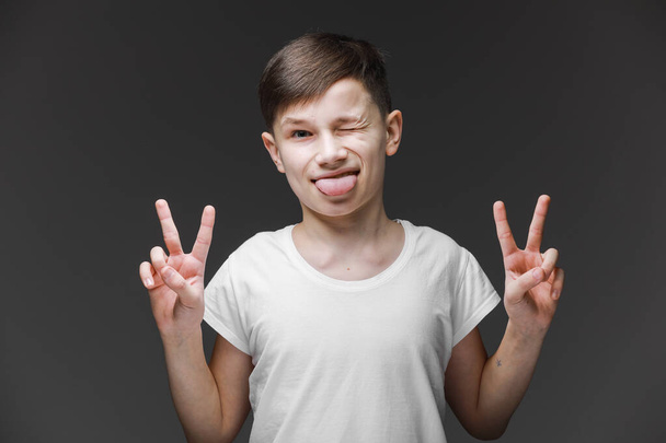 白いTシャツを着た10代の少年が、舌で笑顔で灰色の背景に立っていて、両手の指が勝利サインをしているのを見せている。面白い表現で満足しています。感情の概念第二番 - 写真・画像