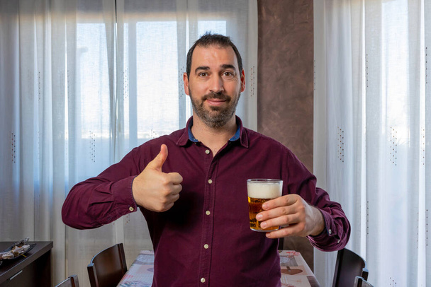 Attraktiver lächelnder bärtiger Mann im lila Hemd mit erhobenem Daumen und einem Bier in der anderen Hand. Glückskonzept - Foto, Bild