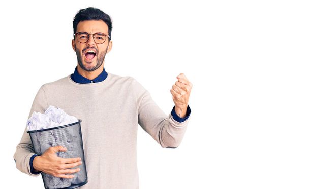 Young handsome hispanic man holding paper bin full of crumpled papers screaming proud, celebrating victory and success very excited with raised arms  - Foto, afbeelding