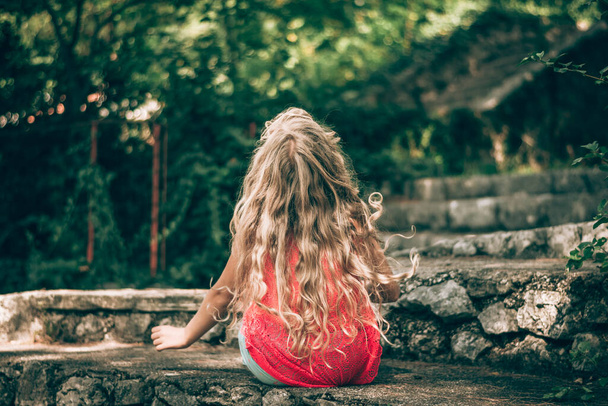 terug uitzicht op jonge tiener meisje met lange krullend blond haar zitten in rotsachtige muur - Foto, afbeelding