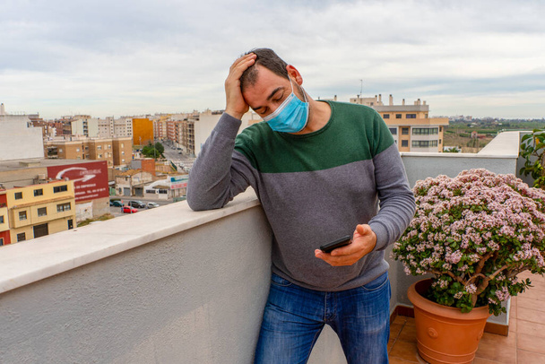 Бородатый мужчина в сером и зеленом свитере в хирургической маске, чтобы защитить себя от коронавируса, с тревогой смотрящего на свой сотовый телефон на террасе своего дома. Технологии и здравоохранение - Фото, изображение