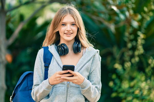 Kaunis valkoihoinen opiskelija teini hymyilee onnellisena älypuhelimella puistossa - Valokuva, kuva
