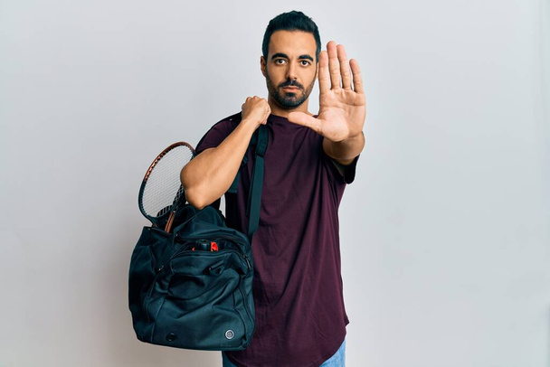 Молодой латиноамериканец держит спортивную сумку с открытой рукой делает знак стоп с серьезным и уверенным выражением, защитный жест  - Фото, изображение
