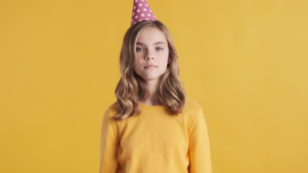 Beautiful blond teenage girl with wavy hair wearing party hat waiting guests on her birthday party over yellow background - Footage, Video