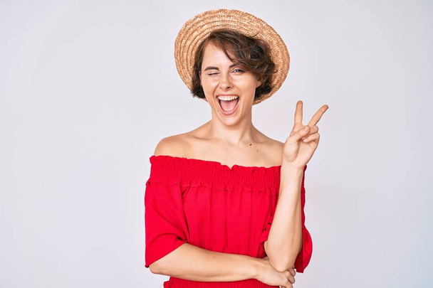 Молода іспаномовна жінка в літньому капелюсі посміхається з щасливим обличчям, підморгнувши на камеру, роблячи знак перемоги. номер два
.  - Фото, зображення
