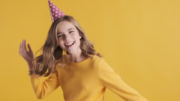 Schöne aufgeregte blonde Teenager-Mädchen mit Geburtstagsmütze sieht glücklich tanzen in der Kamera während der Geburtstagsparty über gelbem Hintergrund. Positives Teenie-Mädchen - Filmmaterial, Video