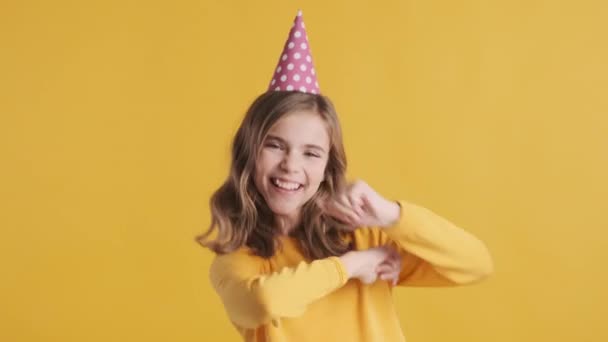 パーティーの帽子をかぶっているかなり陽気なブロンドのティーンエイジャーの女の子は、黄色の背景の上に誕生日に踊ります。表現の周りをうろうろ - 映像、動画