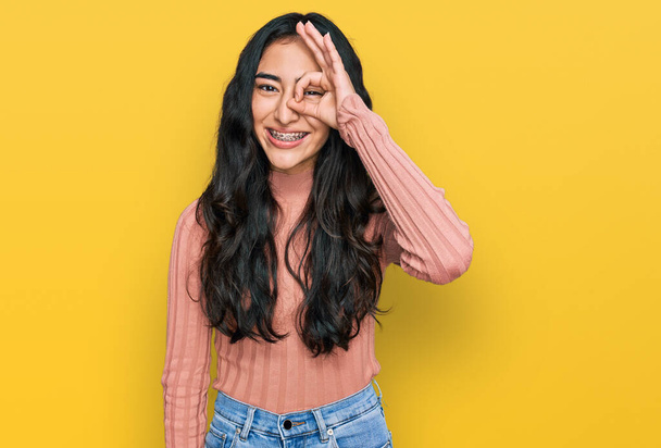 Hispanisches Teenager-Mädchen mit Zahnspange in lässiger Kleidung tut ok Geste mit lächelnden Händen, Augen durch Finger mit glücklichem Gesicht.  - Foto, Bild