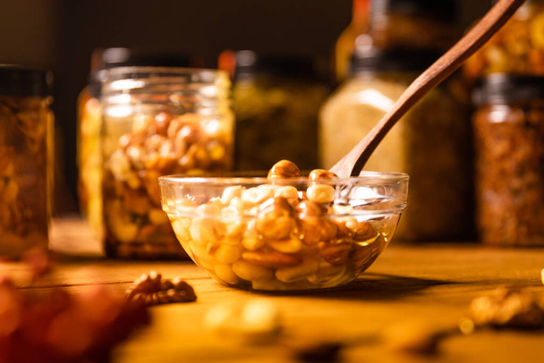 Honey mixed with nuts, seeds in a transparent plate with wooden spoon, one jar, walnuts kernel, red rowan, on rustic wooden kitchen table. Still life photography. - Photo, Image