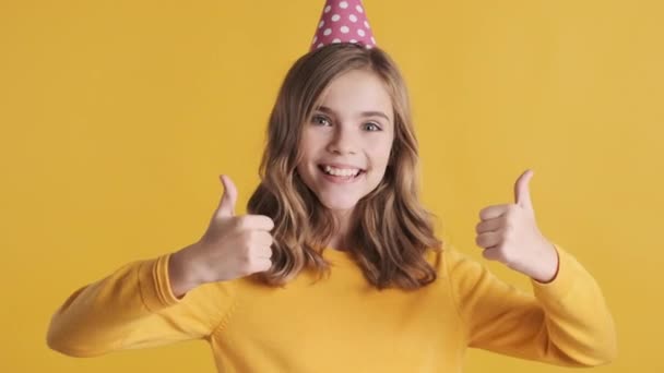 Chica adolescente rubia bastante positiva con sombrero de fiesta que se ve feliz manteniendo los pulgares hacia arriba en la cámara sobre el fondo amarillo. Chica alegre como su fiesta de cumpleaños - Metraje, vídeo