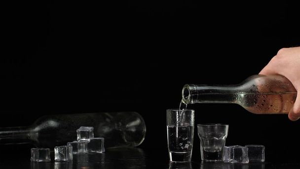 Barman gieten van twee shots van wodka met ijsblokjes uit fles in glazen op zwarte achtergrond - Foto, afbeelding